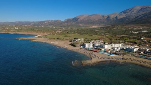 Free Scenic aerial view of Malia, Crete featuring a beachfront resort, clear blue sea, and mountainous landscape. Stock Photo