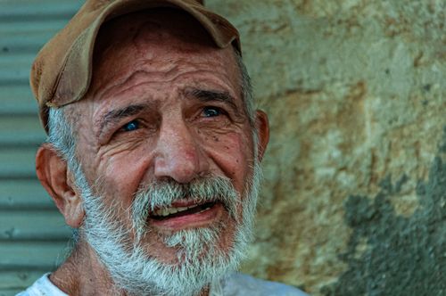 Free Portrait of an elderly man wearing a cap, smiling thoughtfully against a textured outdoor wall. Stock Photo