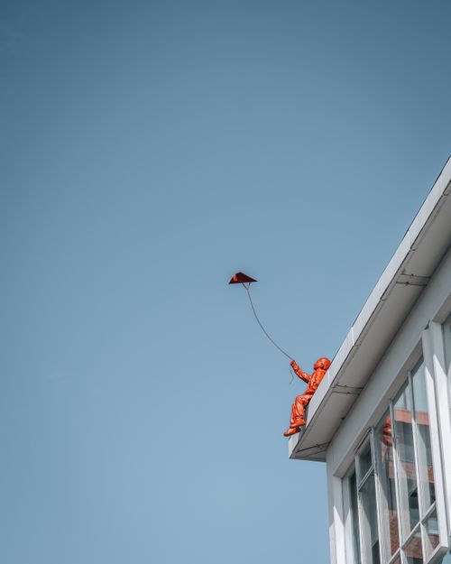Free Vibrant red sculpture of a child flying a kite on a building rooftop against a clear blue sky in Amsterdam. Stock Photo