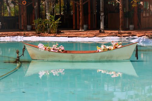 Free A small boat decorated with flowers floats peacefully in a serene blue pool beside wooden buildings. Stock Photo