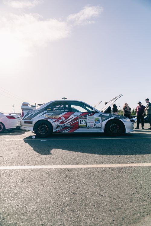 Free Side view of a race car with its hood raised under a clear blue sky, surrounded by spectators. Stock Photo