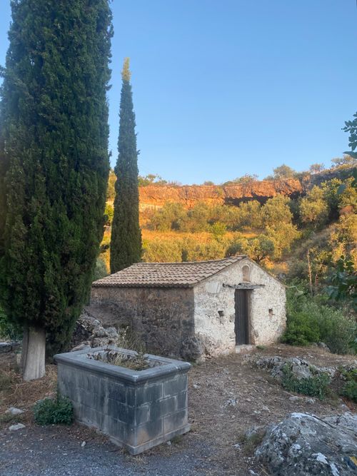 Free A rustic stone chapel surrounded by lush greenery and cypress trees in the Greek countryside at sunset. Stock Photo