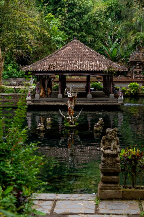 Free Serene Balinese pavilion surrounded by lush greenery and calm waters, featuring traditional statues. Stock Photo