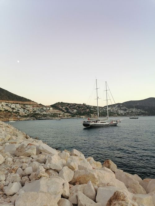 Free A serene sailing boat anchors in a calm Aegean bay at twilight, with rocky shores and hillside houses in view. Stock Photo