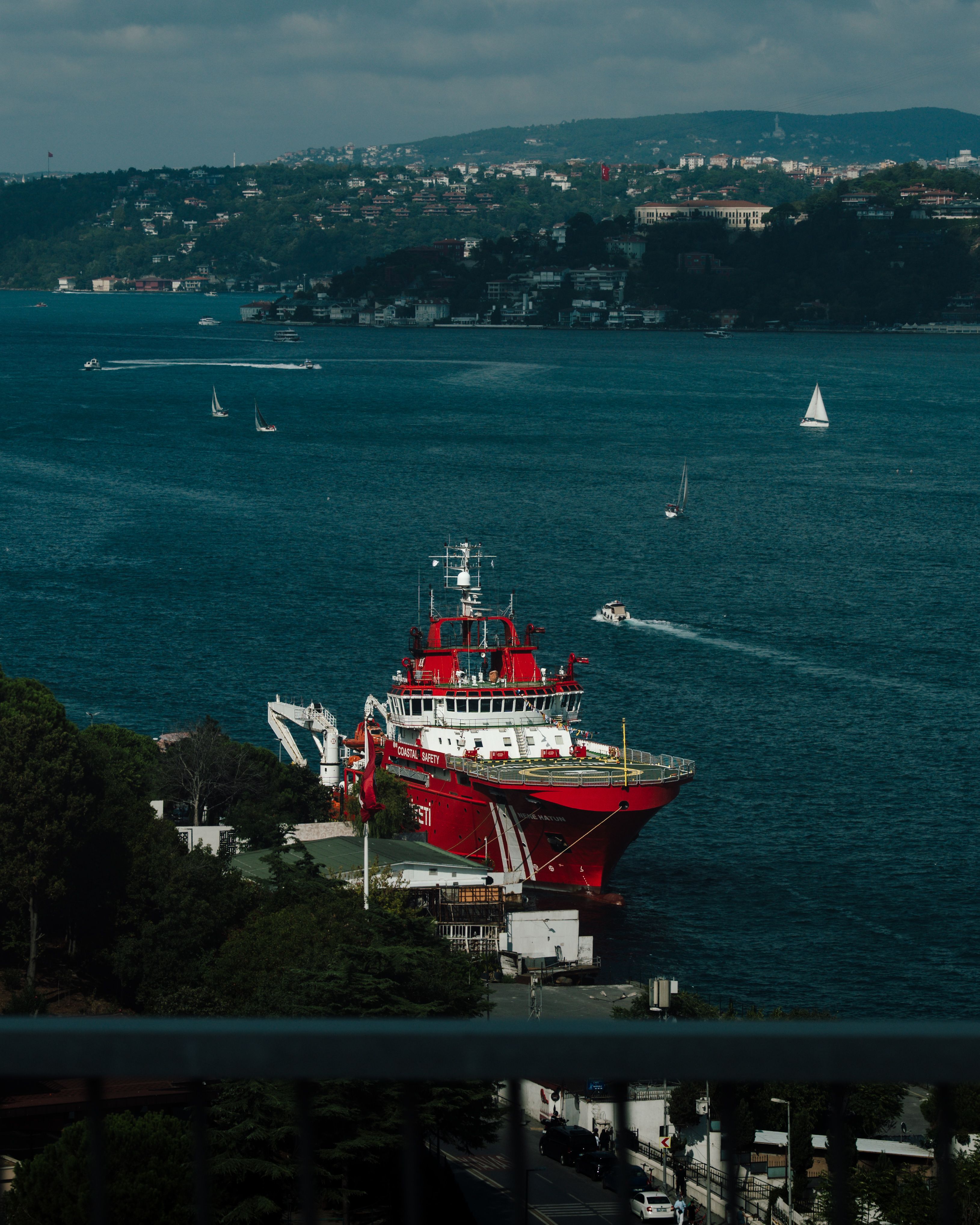 A striking red ship docked at a picturesque harbor with surrounding lush green hills and sailing boats.