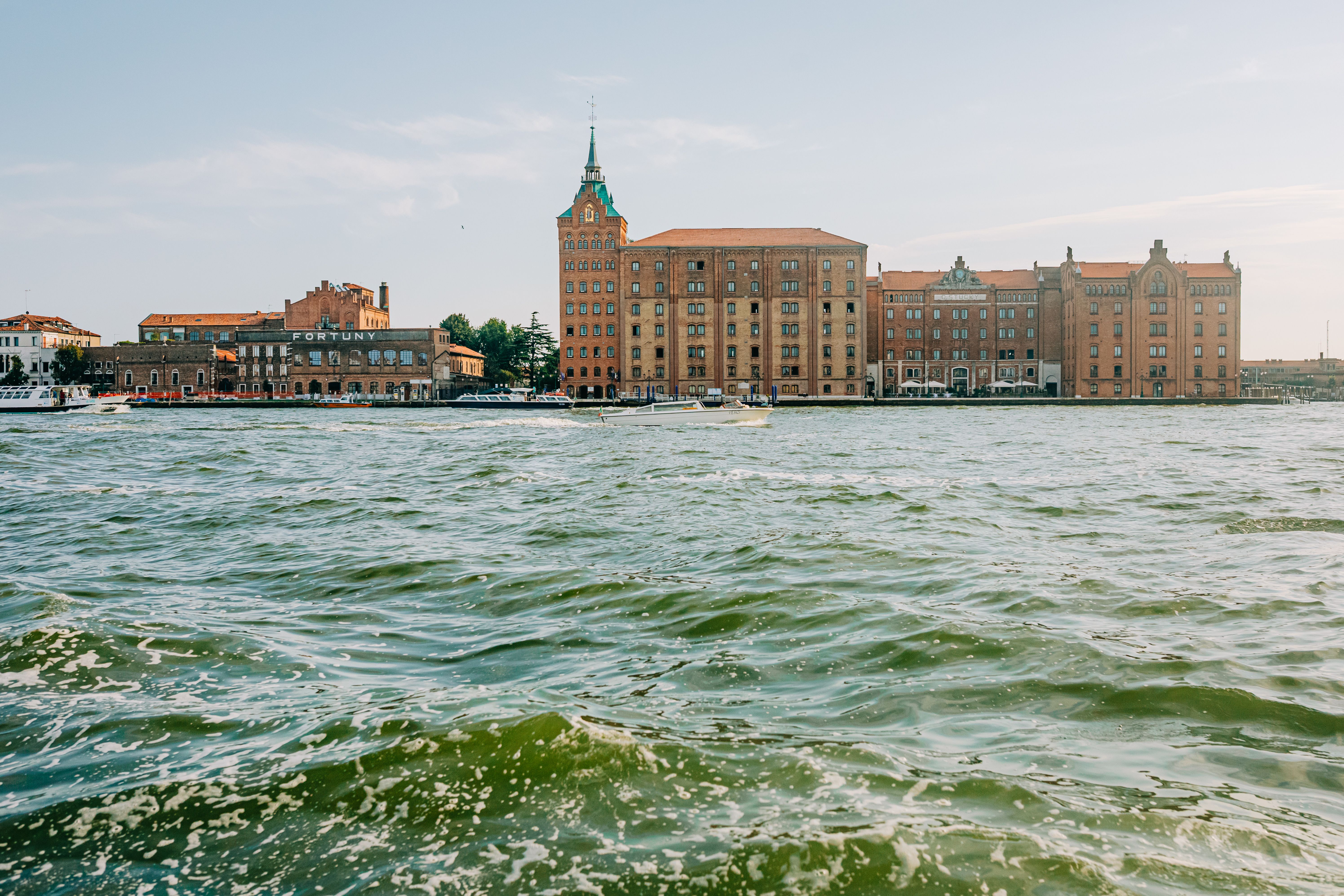 Historic buildings along Venice's waterfront under a sunny sky, reflecting in vivid green waters.