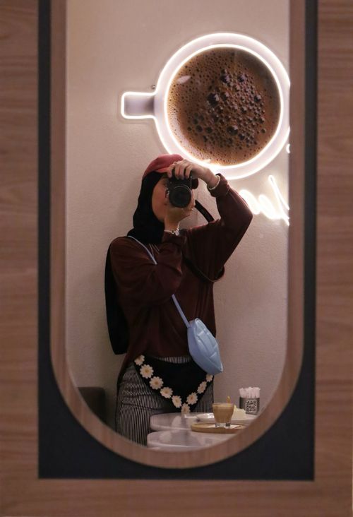 Free A creative mirror selfie in a cafe featuring a glowing neon coffee cup wall art. Stock Photo