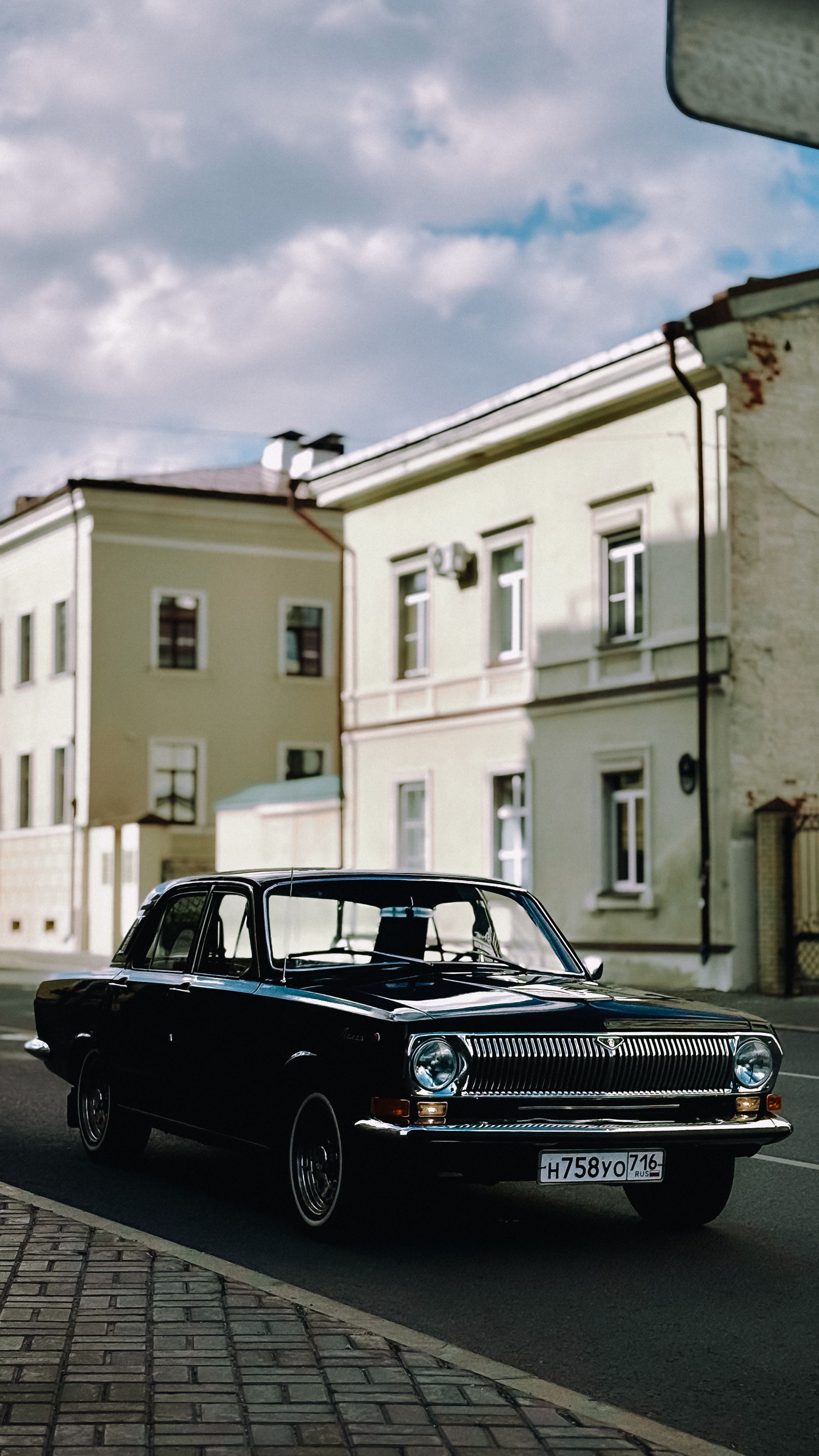 Classic black vintage car driving past historic buildings under a cloudy sky.