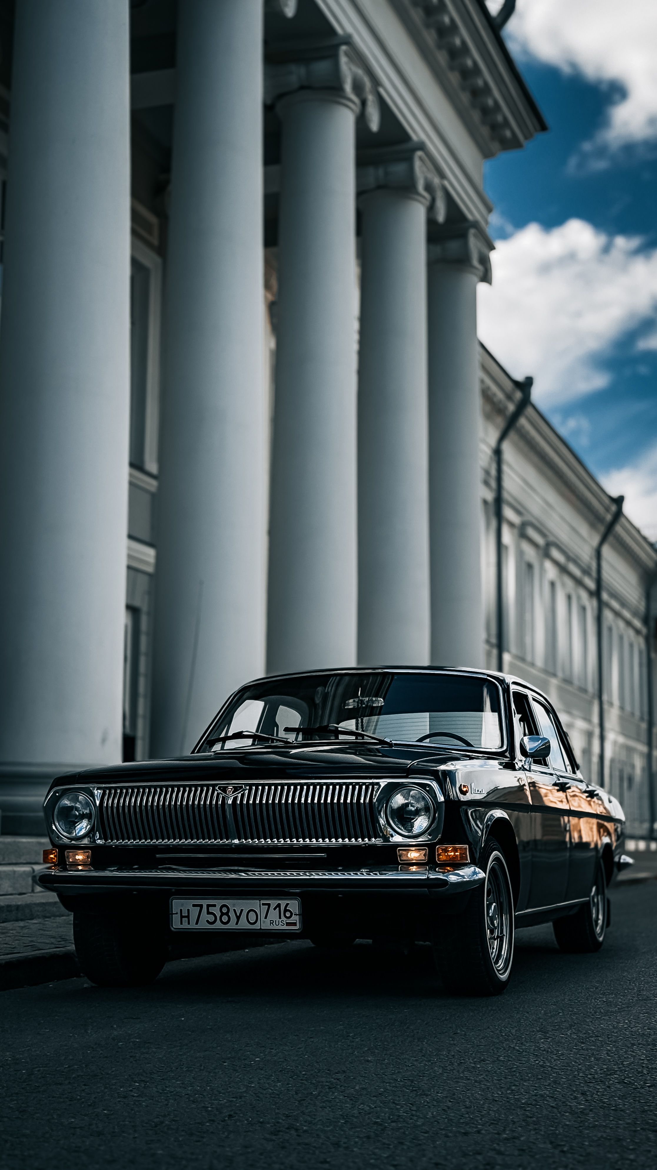 A striking image of a vintage black car parked in front of majestic white columns under a bright blue sky.