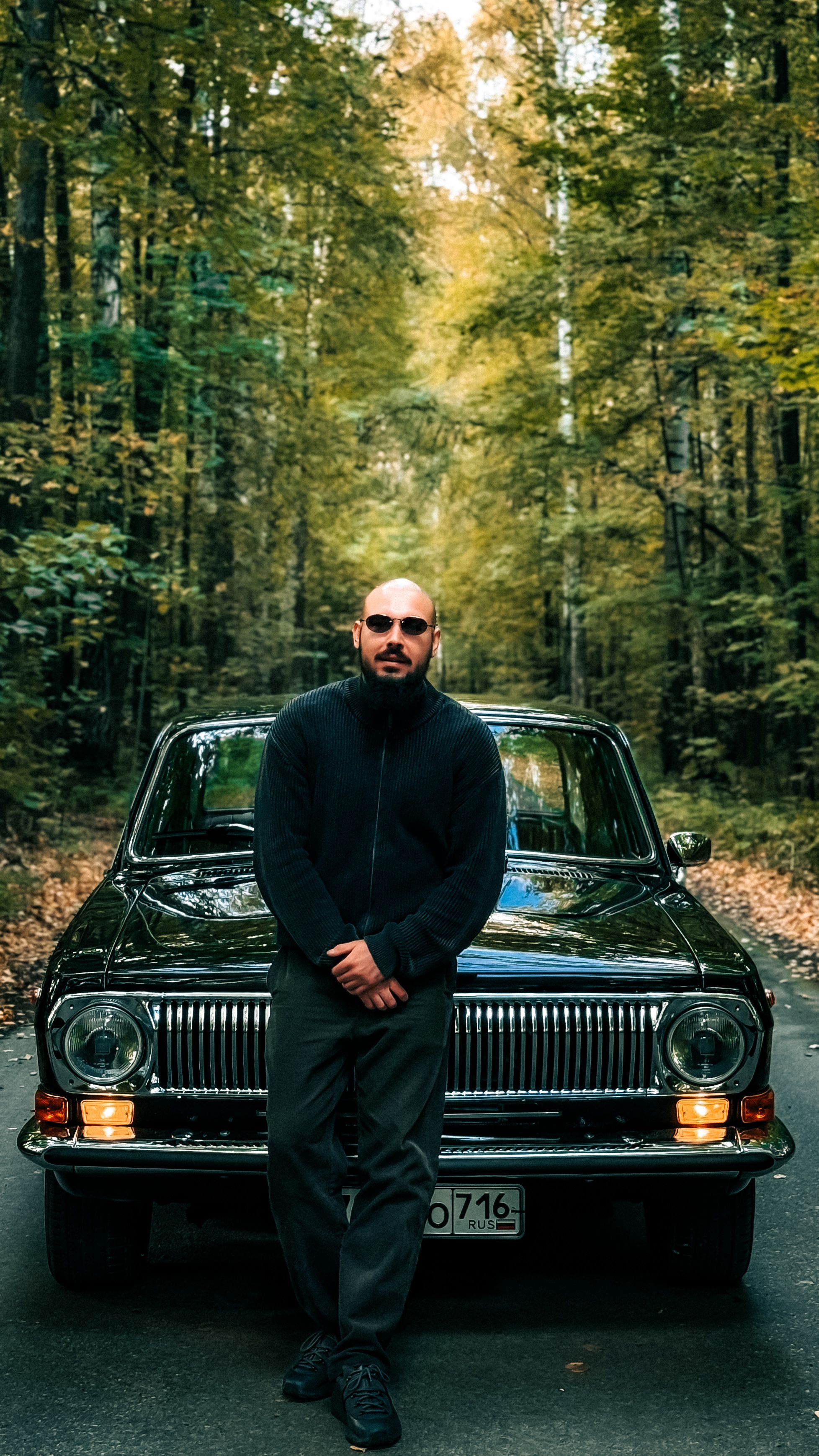 A man poses confidently in front of a classic car surrounded by vibrant autumn trees.