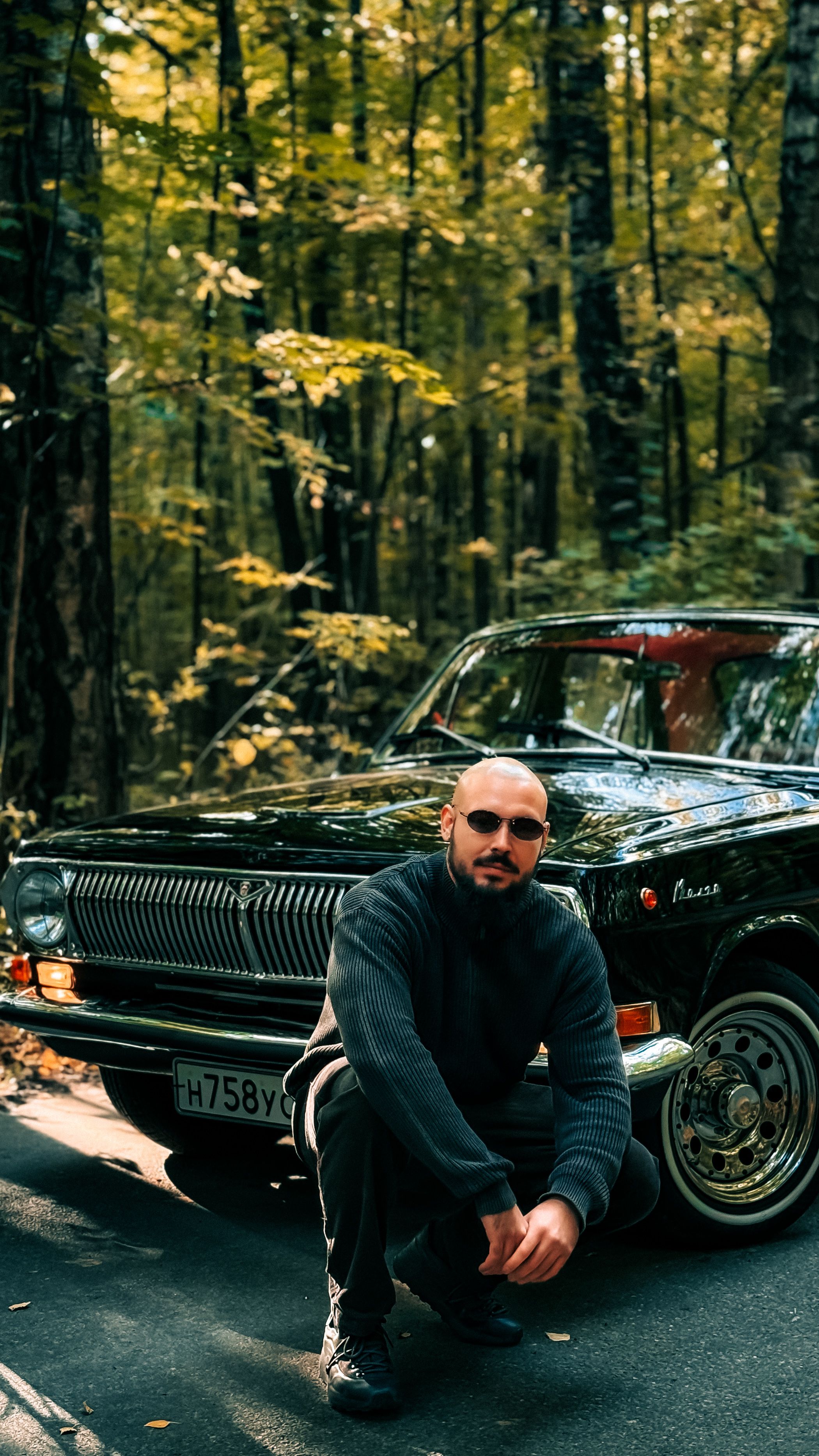 Man in a forest kneels by a vintage black car, surrounded by autumn trees, creating a serene and nostalgic scene.