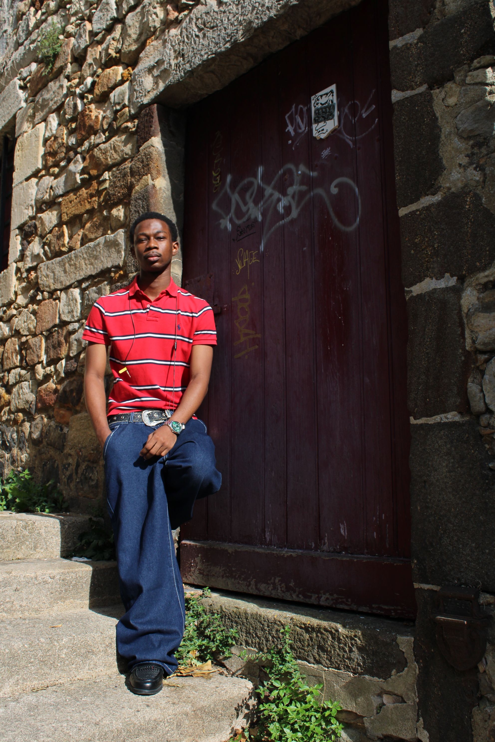 Young man in red shirt sitting on stone steps outside rustic building.