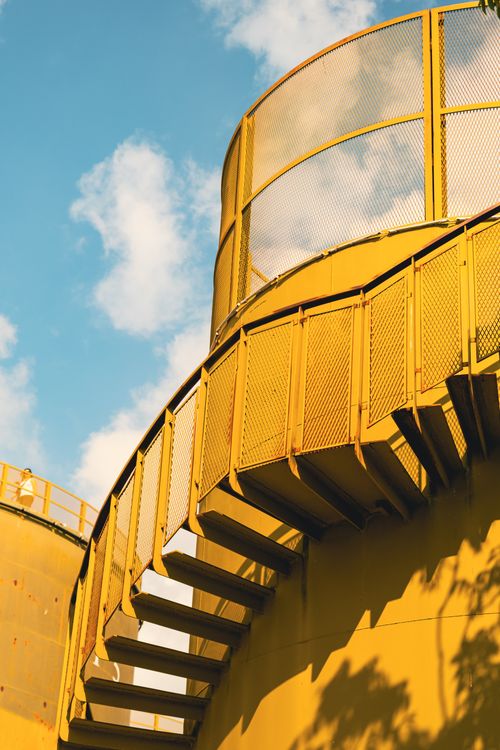 Free A vibrant yellow industrial staircase with mesh railing set against a clear blue sky. Stock Photo