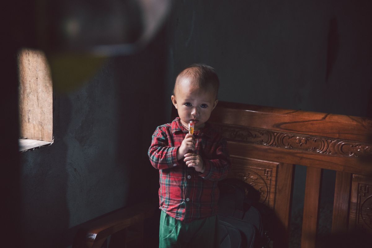 A young child in casual clothing holding a candy indoors, illuminated by natural window light.