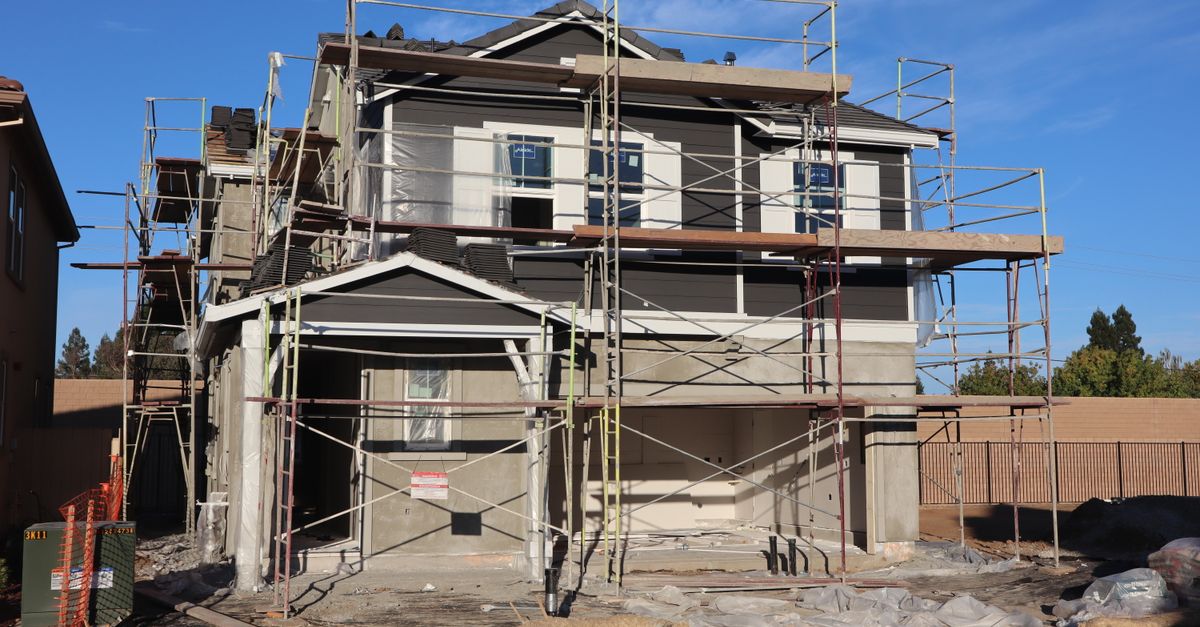 A two-story house under construction, surrounded by scaffolding on a clear, sunny day.