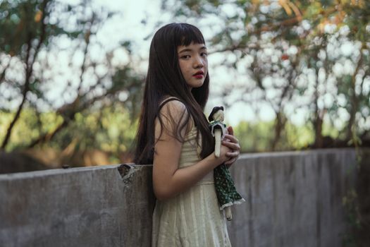 一个年轻女孩抱着洋娃娃，站在绿树成荫的户外，陷入沉思