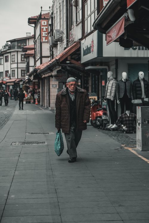Free An elderly man walks through a city street lined with shops, showing urban life and culture. Stock Photo