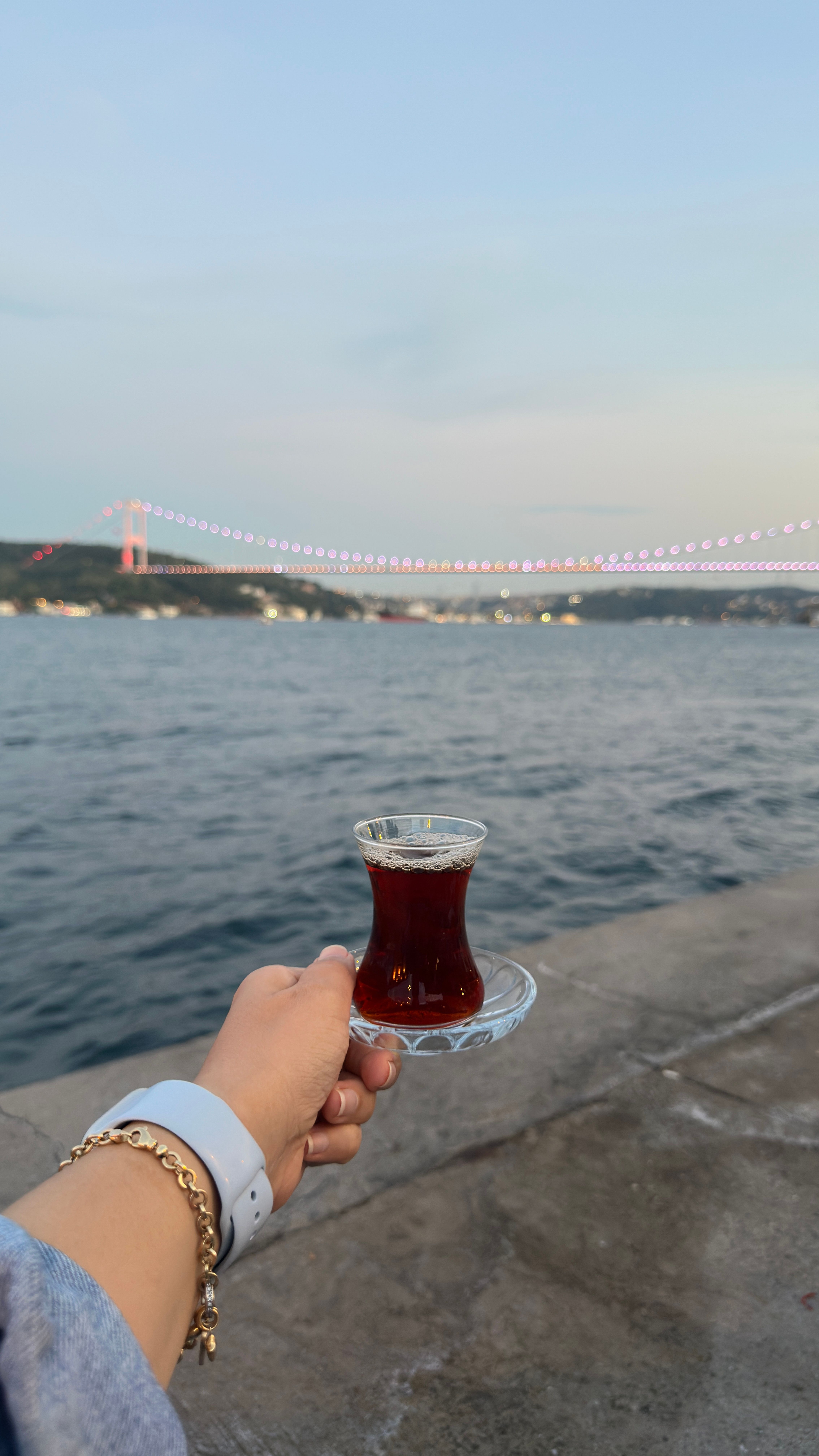 A hand holding a traditional Turkish tea with the Bosphorus Bridge in the background at twilight.