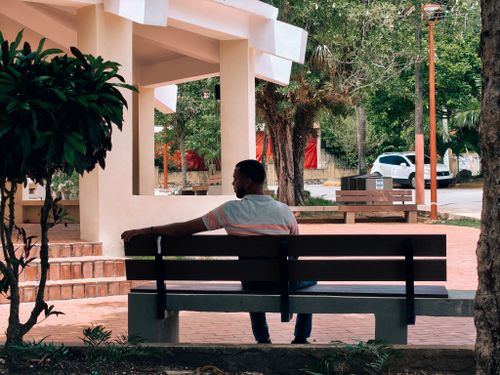 Free A man sits on a bench in a peaceful urban park surrounded by greenery and architecture. Stock Photo