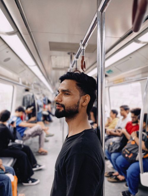 Free Profile view of a young man standing inside a bustling metro train with passengers seated around. Stock Photo