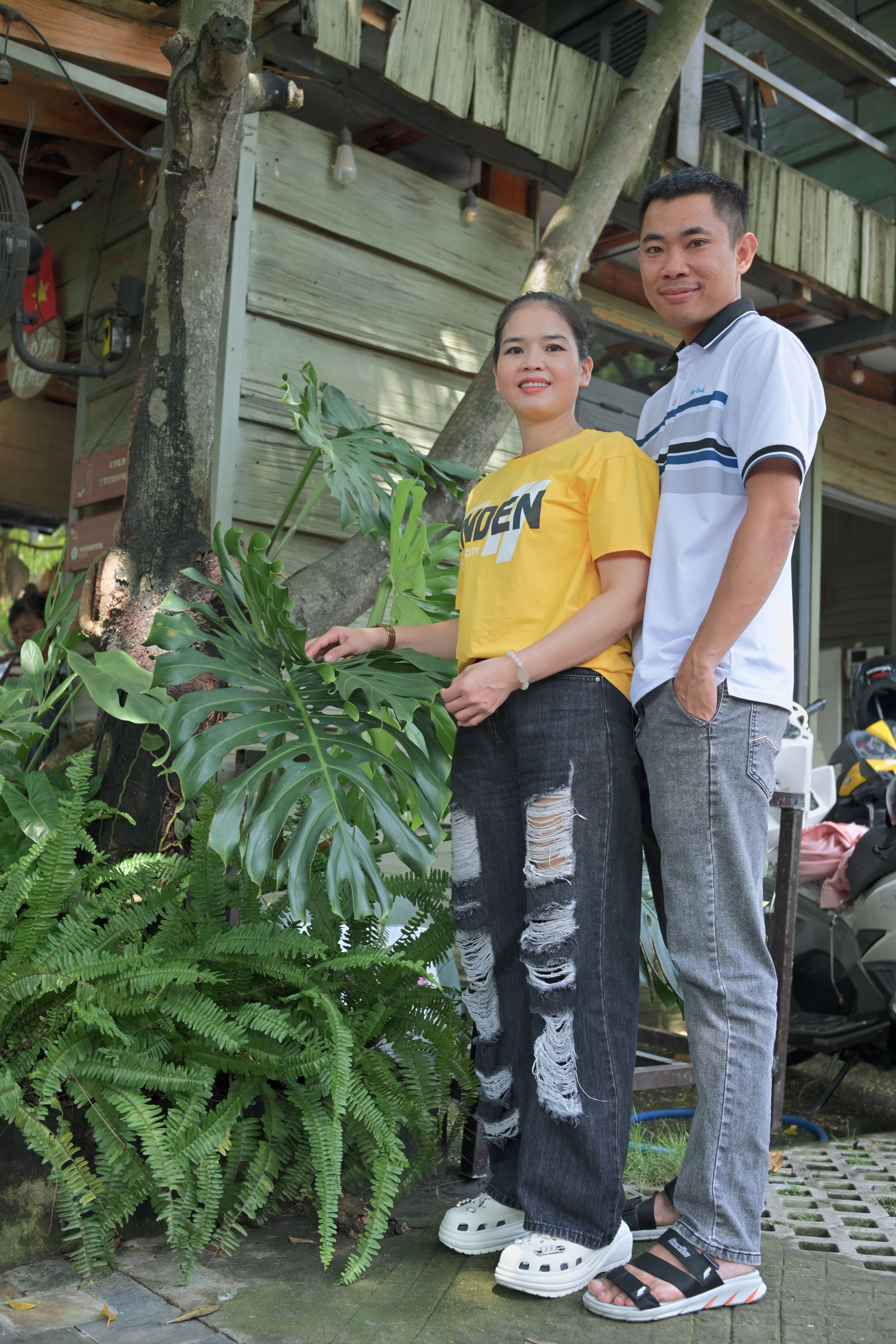 A smiling couple stands by vibrant green plants in an outdoor rustic setting.