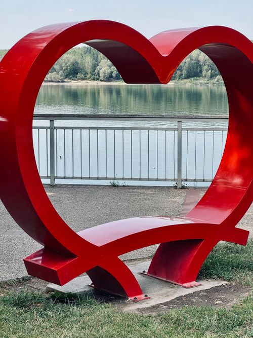 Free A vibrant red heart-shaped bench with a lakeside view, perfect for romantic spots. Stock Photo