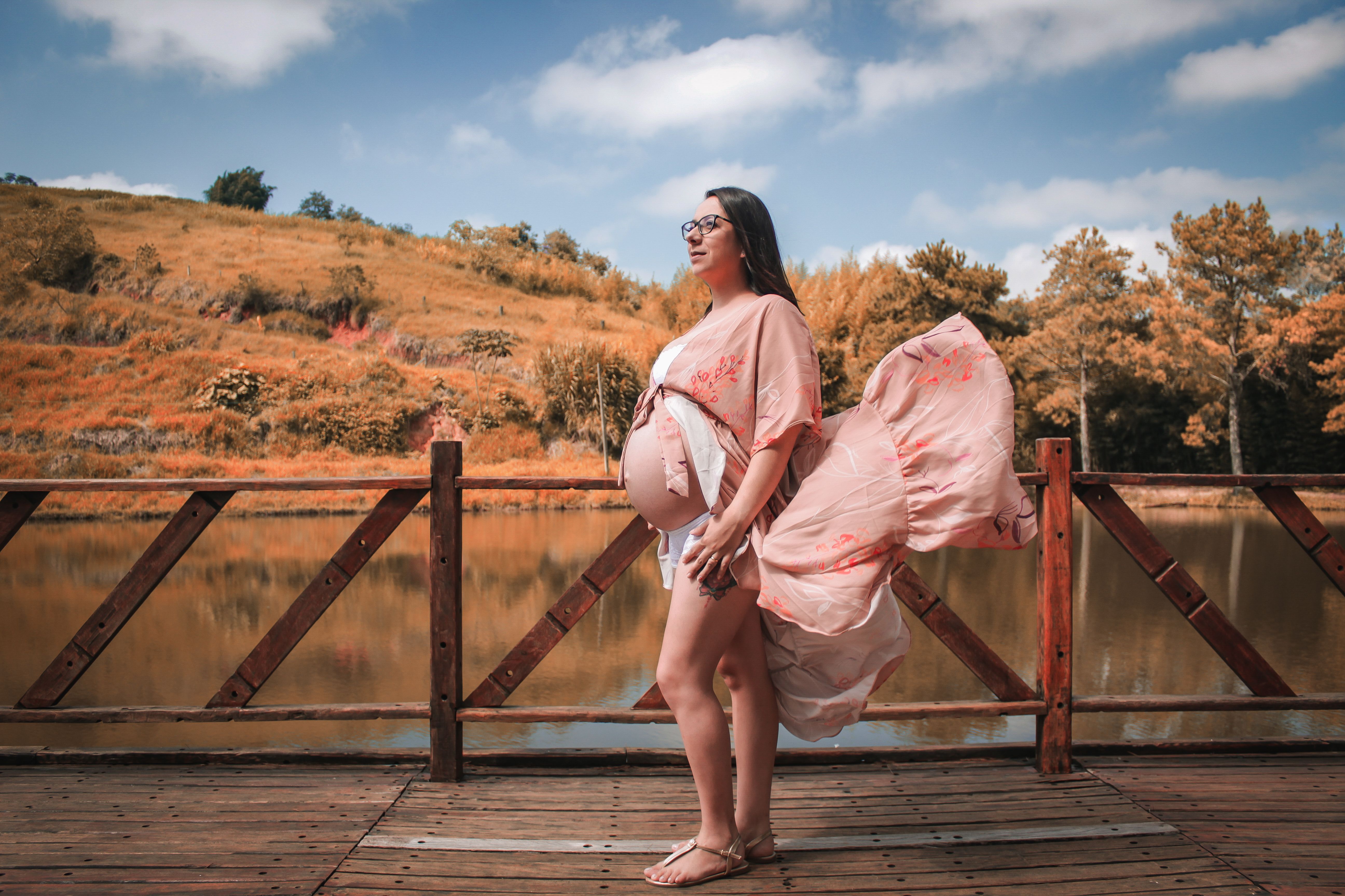 Pregnant woman standing on a bridge by a scenic lake, wearing a flowing dress.