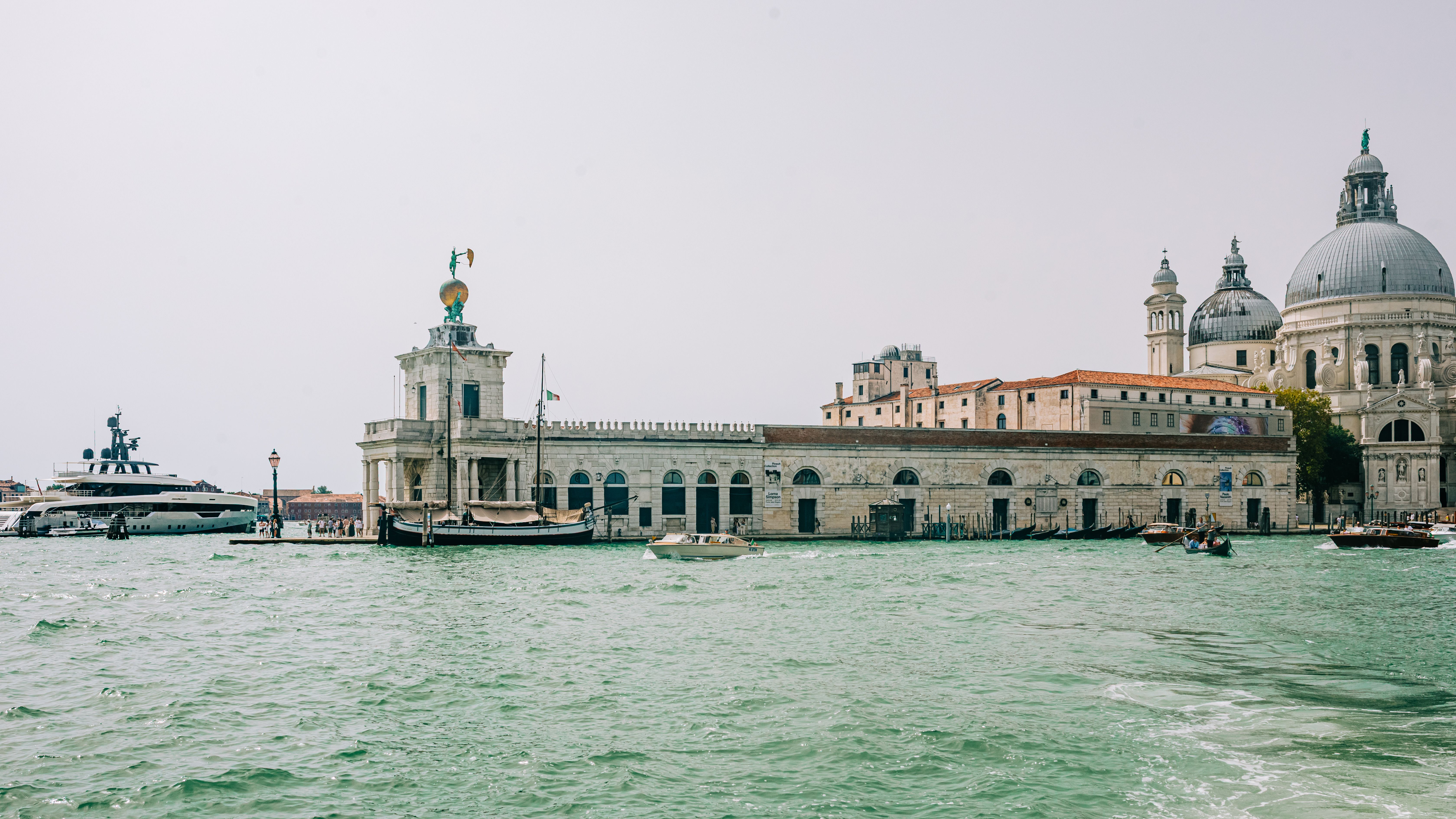A long low building with a small tower and a large domed church beside it on the far side of open green water, with boats moored along the quay