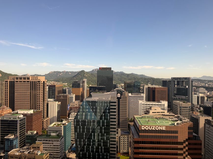 Aerial view of Seoul's cityscape with modern skyscrapers and distant mountains under a clear blue sky.