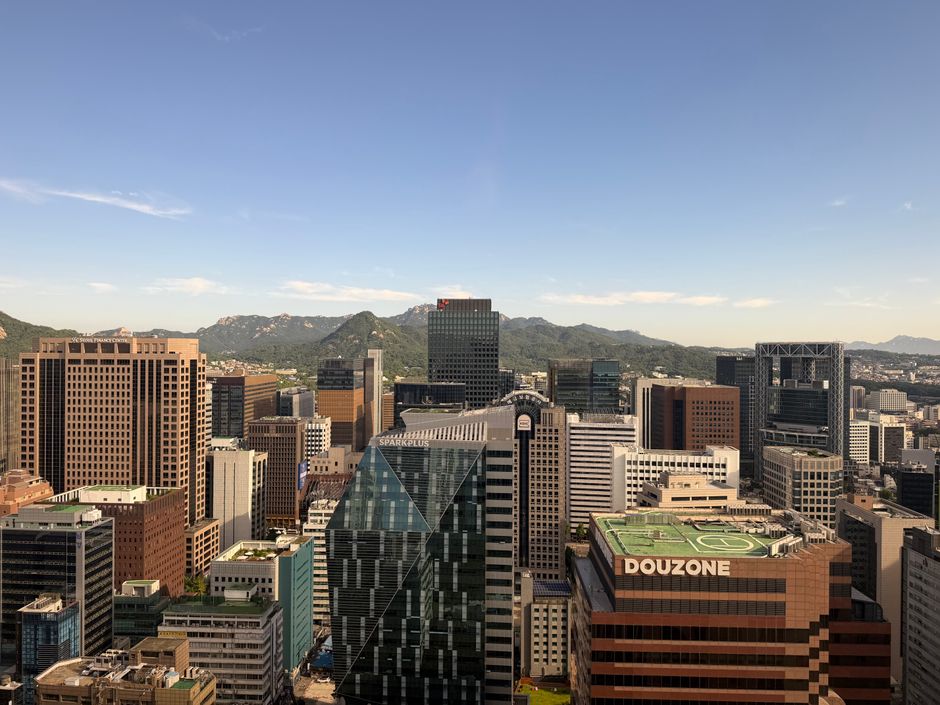 Aerial view of Seoul's cityscape with modern skyscrapers and distant mountains under a clear blue sky.