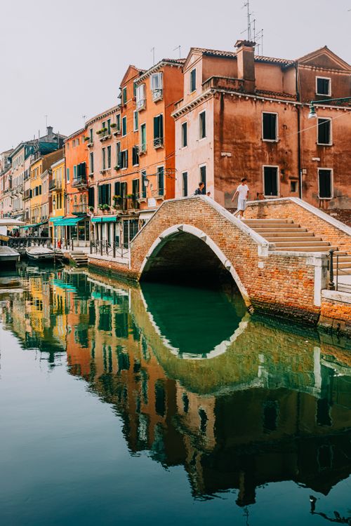 Free Picturesque canal in Venice with colorful historic buildings and a classic arched bridge reflecting in the calm water. Stock Photo