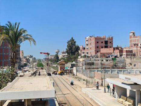 Urban railway station with tracks, buildings, and people under clear blue sky.