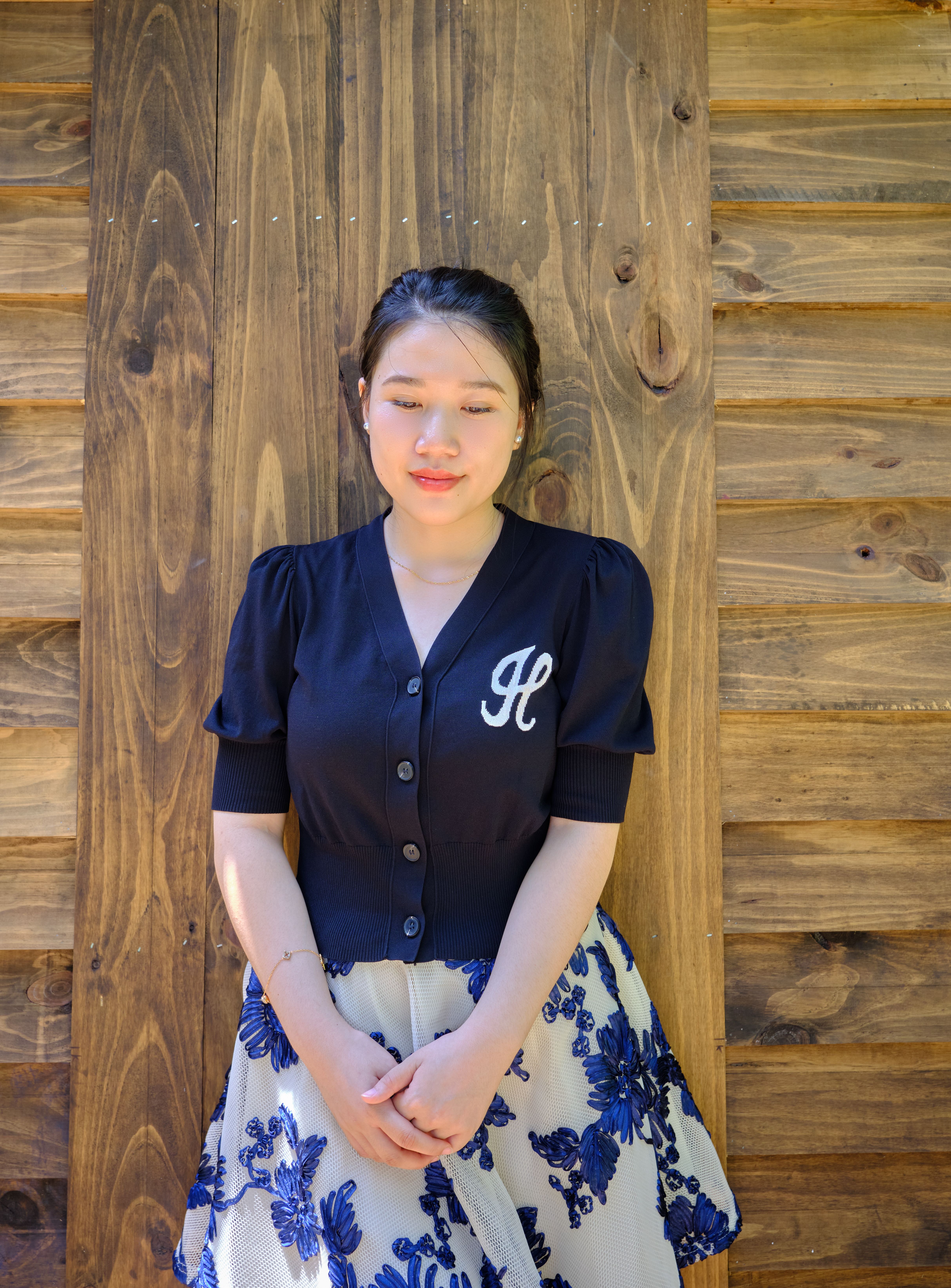 Portrait of a young woman in a floral skirt standing against a wooden wall.