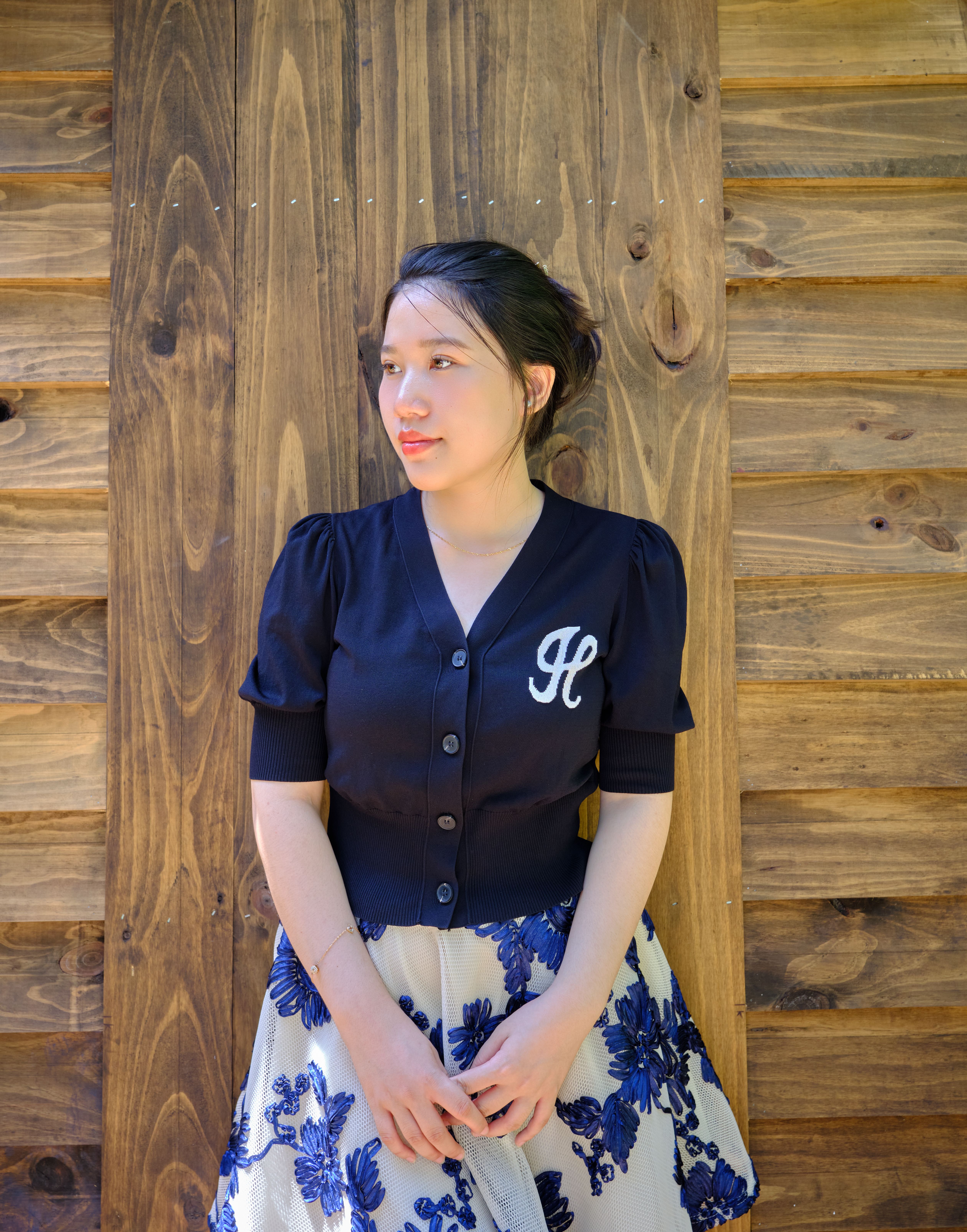 Elegant woman in floral skirt and black blouse posing against a rustic wooden wall.