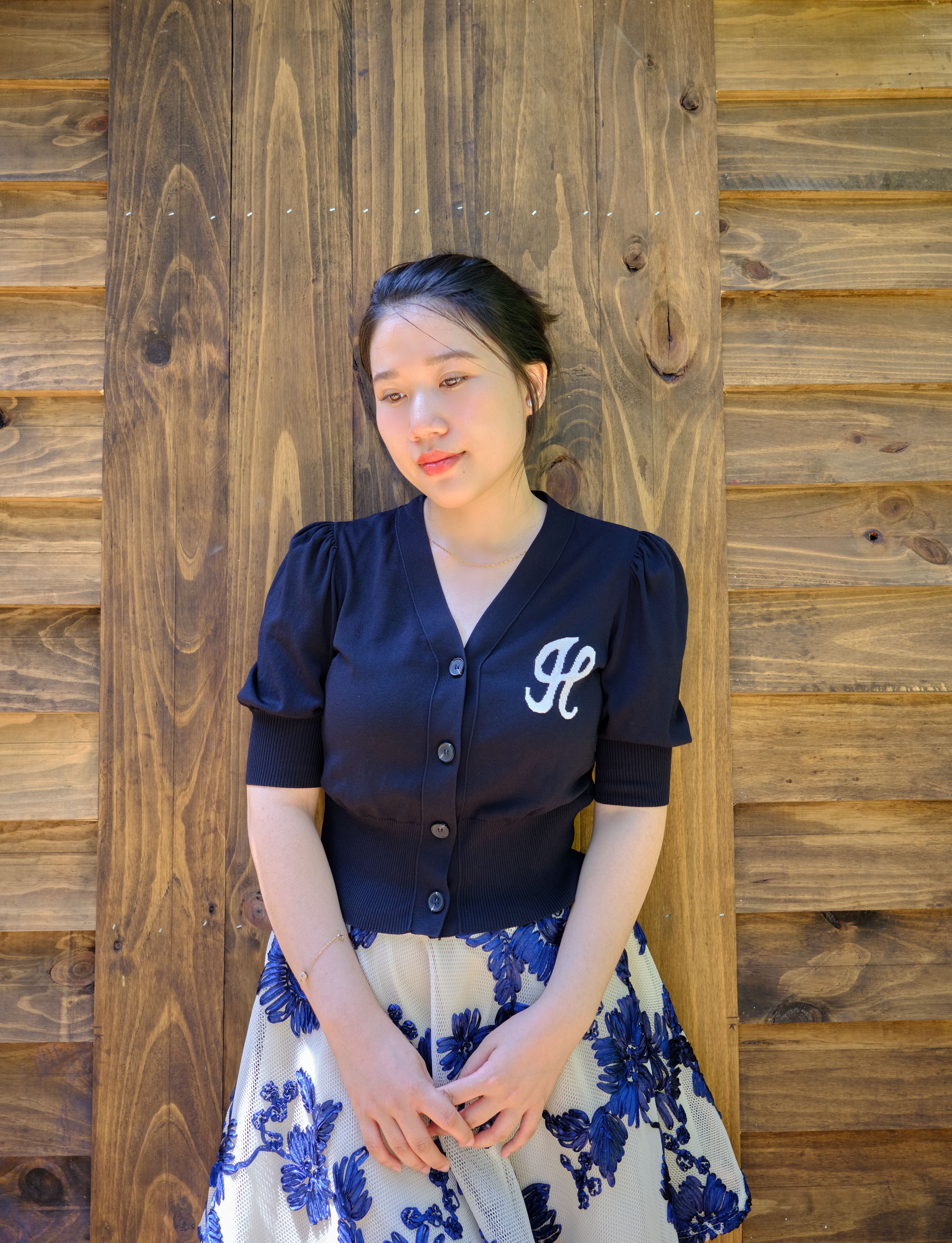 A young woman in a navy cardigan and floral skirt stands by a wooden wall, showcasing modern fashion.