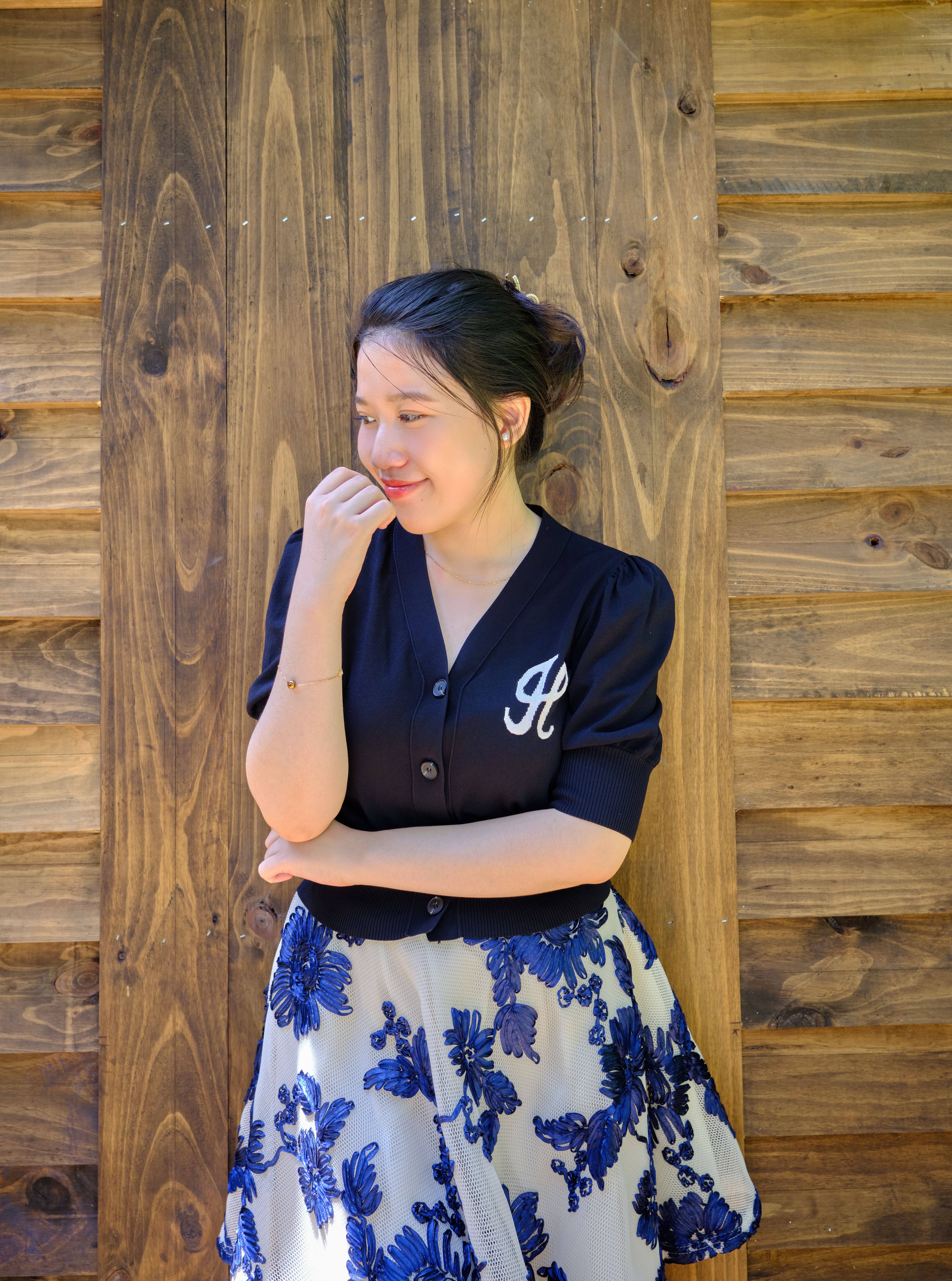 A young woman in a floral skirt smiles thoughtfully against a rustic wooden background.
