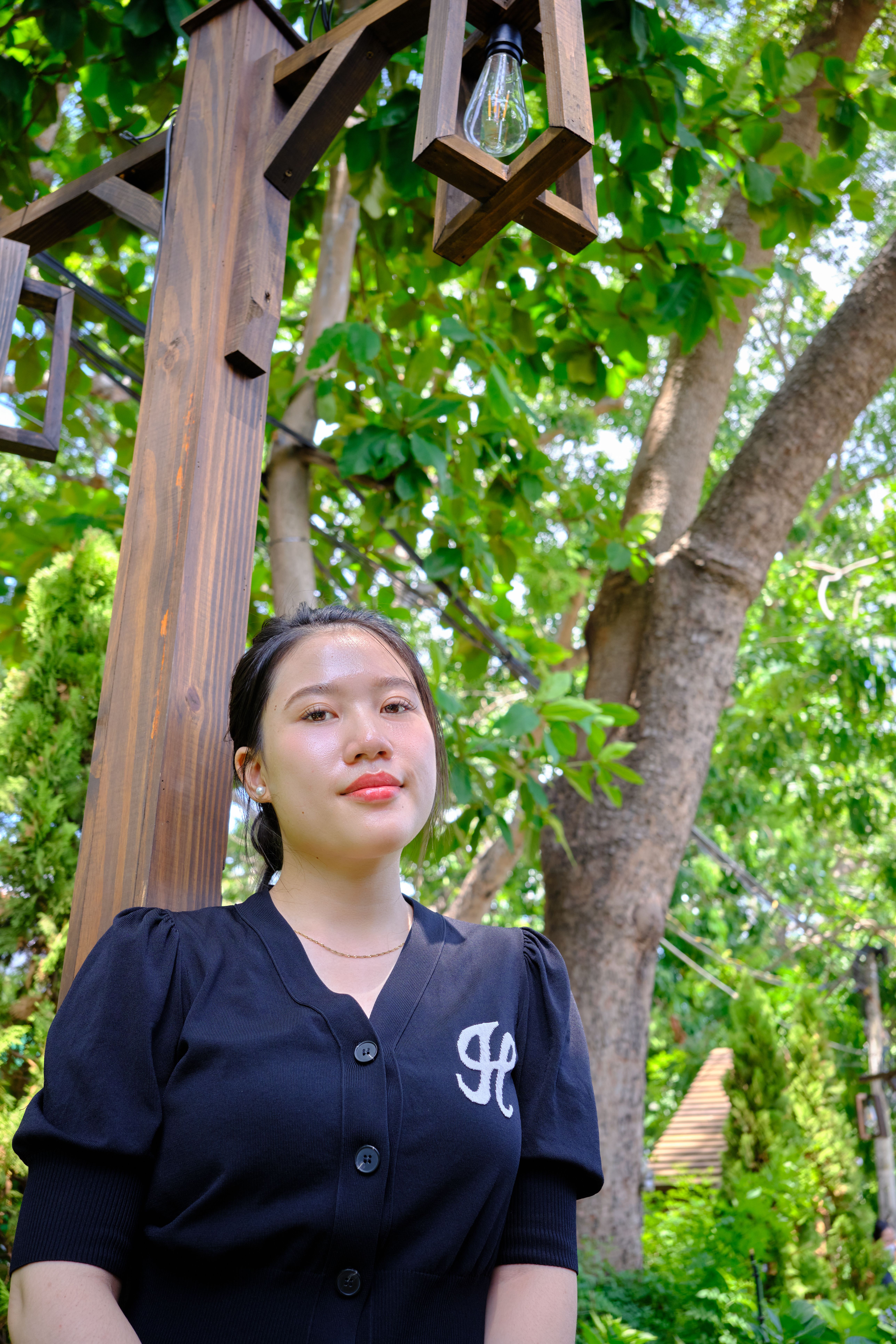 Portrait of a young woman standing under a wooden lamp post surrounded by green trees.