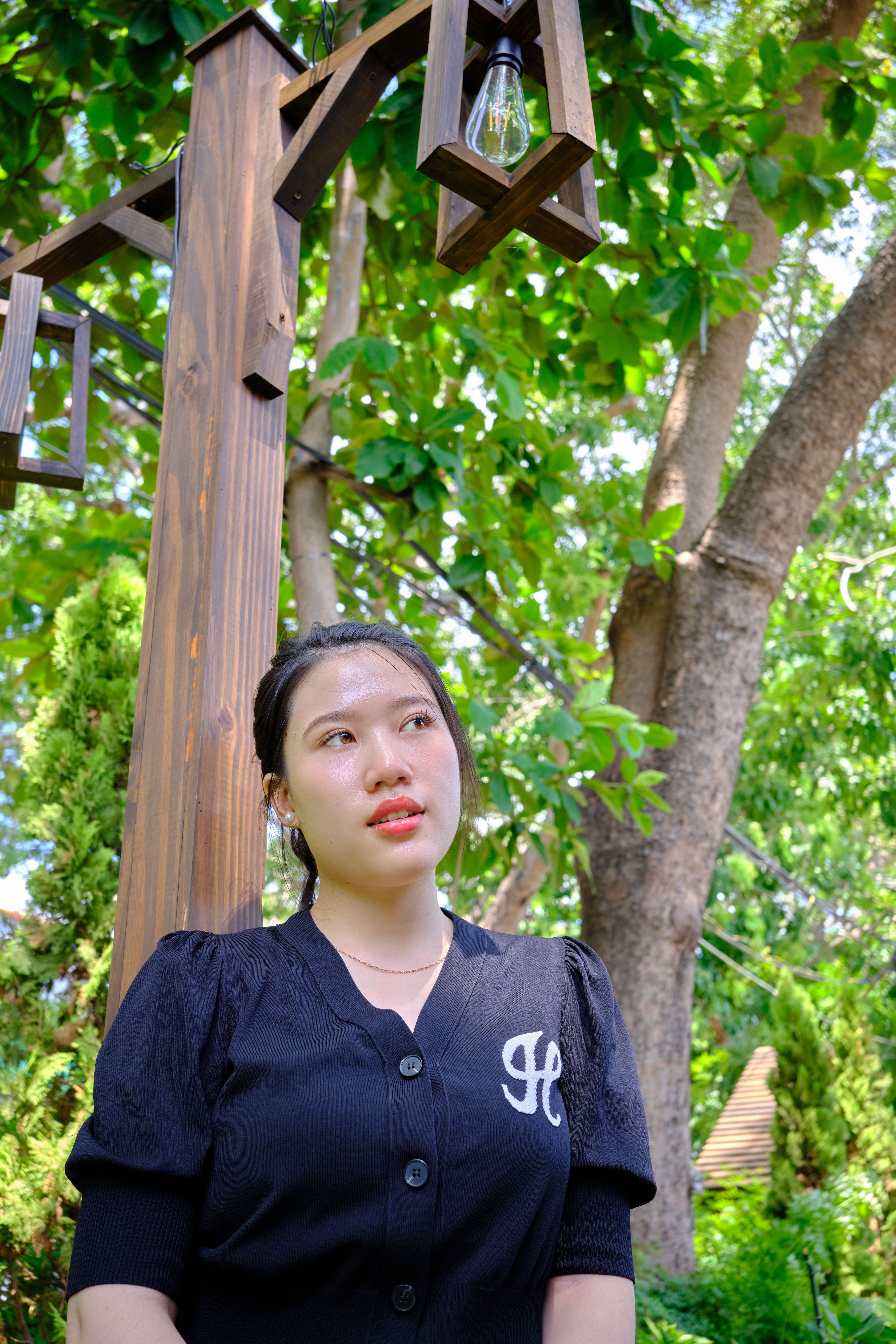 A young woman stands beside a wooden lantern in a lush green setting.