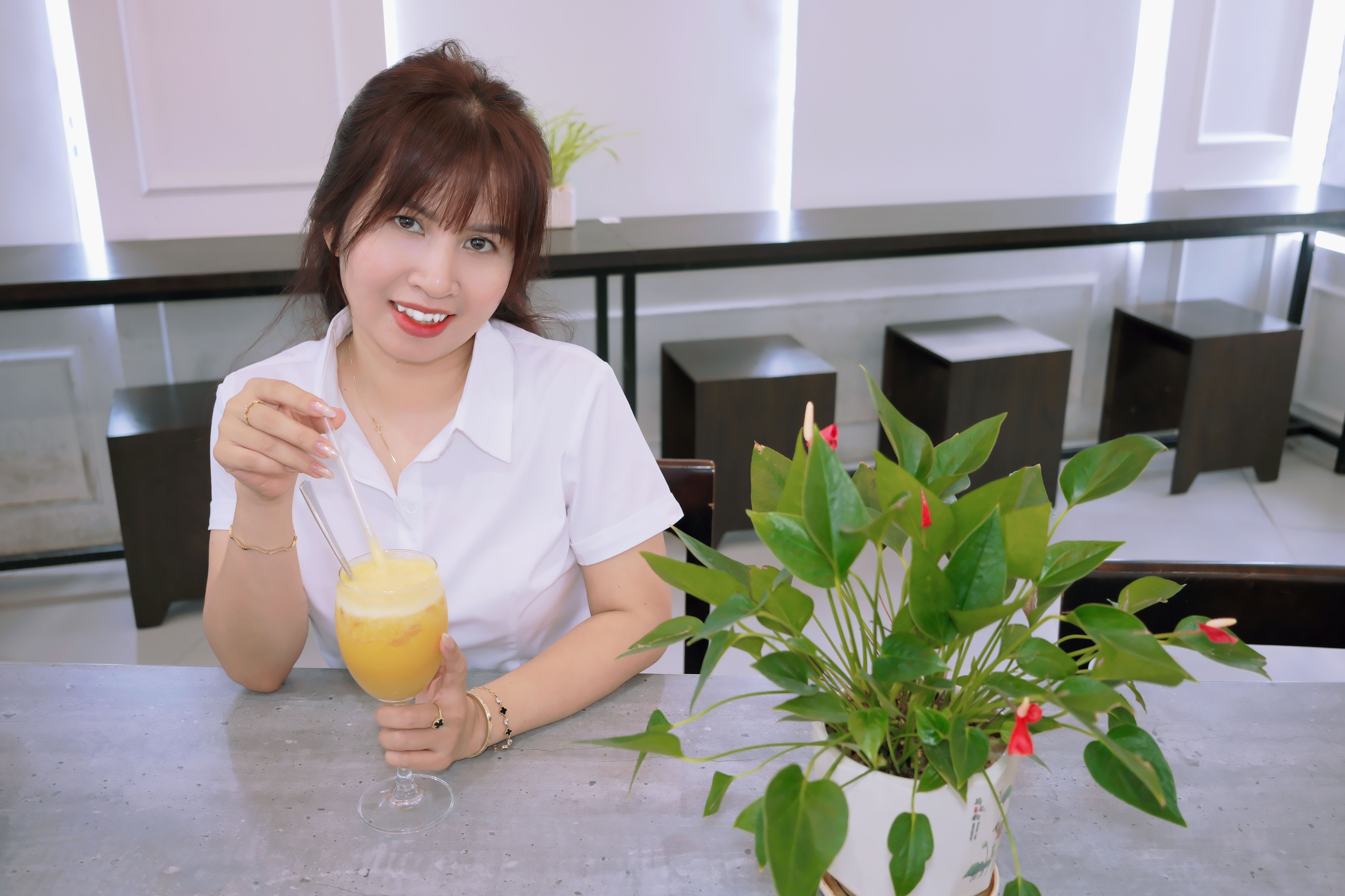 A cheerful woman enjoying a refreshing yellow drink indoors beside a green plant.
