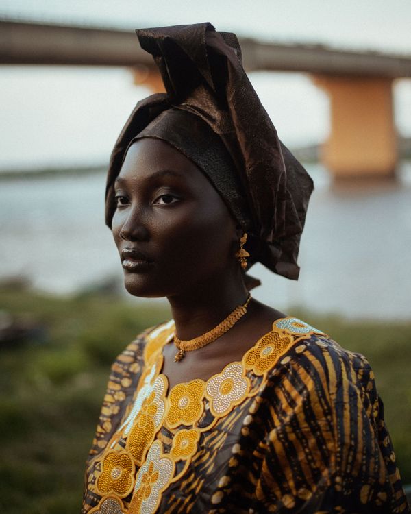 Serene outdoor portrait of an African woman wearing traditional attire and headwrap by a river.