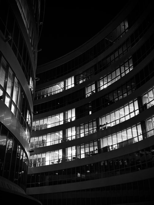 Free Black and white night photograph of a modern office building with illuminated windows showing activity inside. Stock Photo