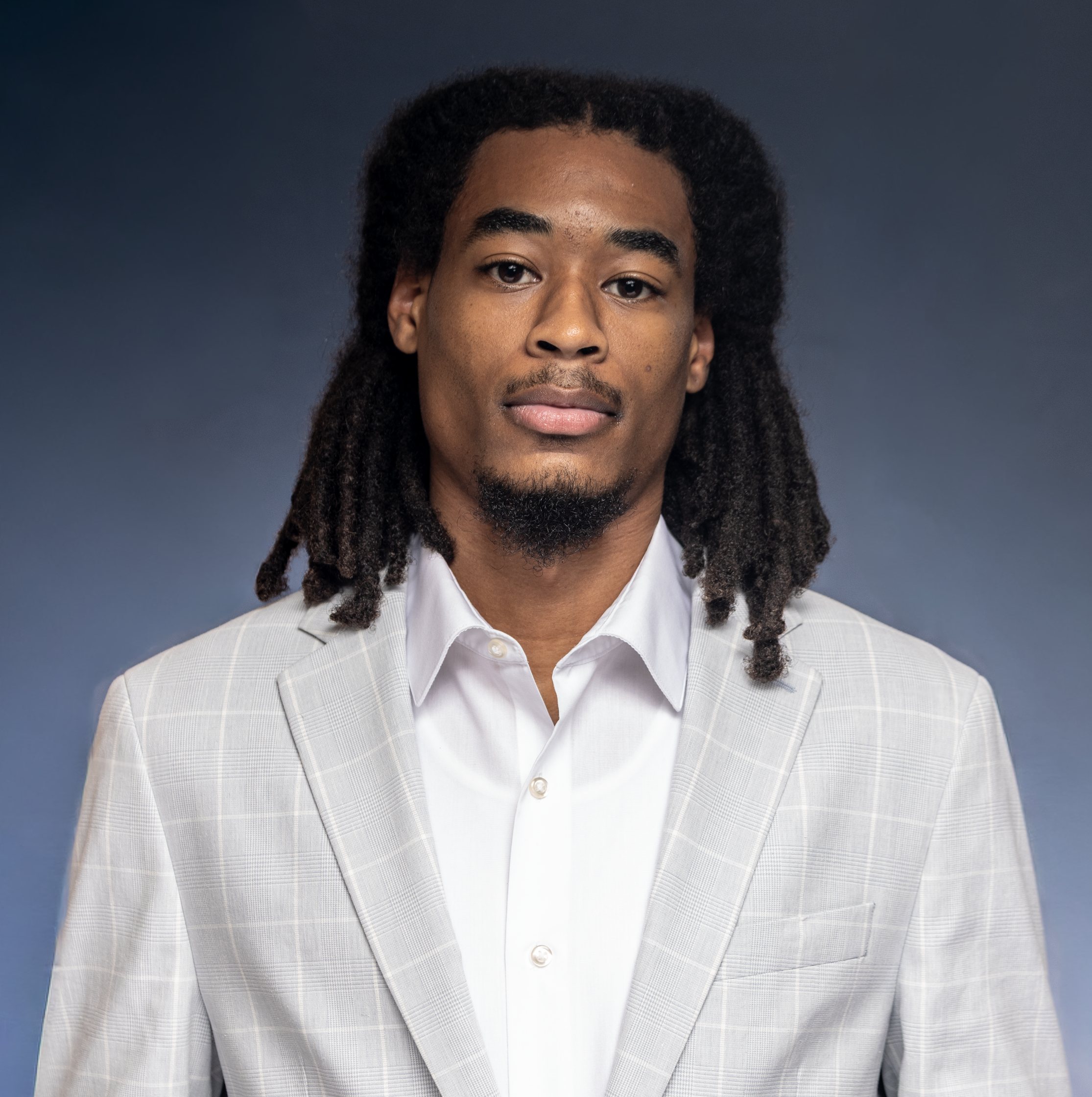 Confident man with dreadlocks wearing a grey plaid suit and white shirt, neutral background.