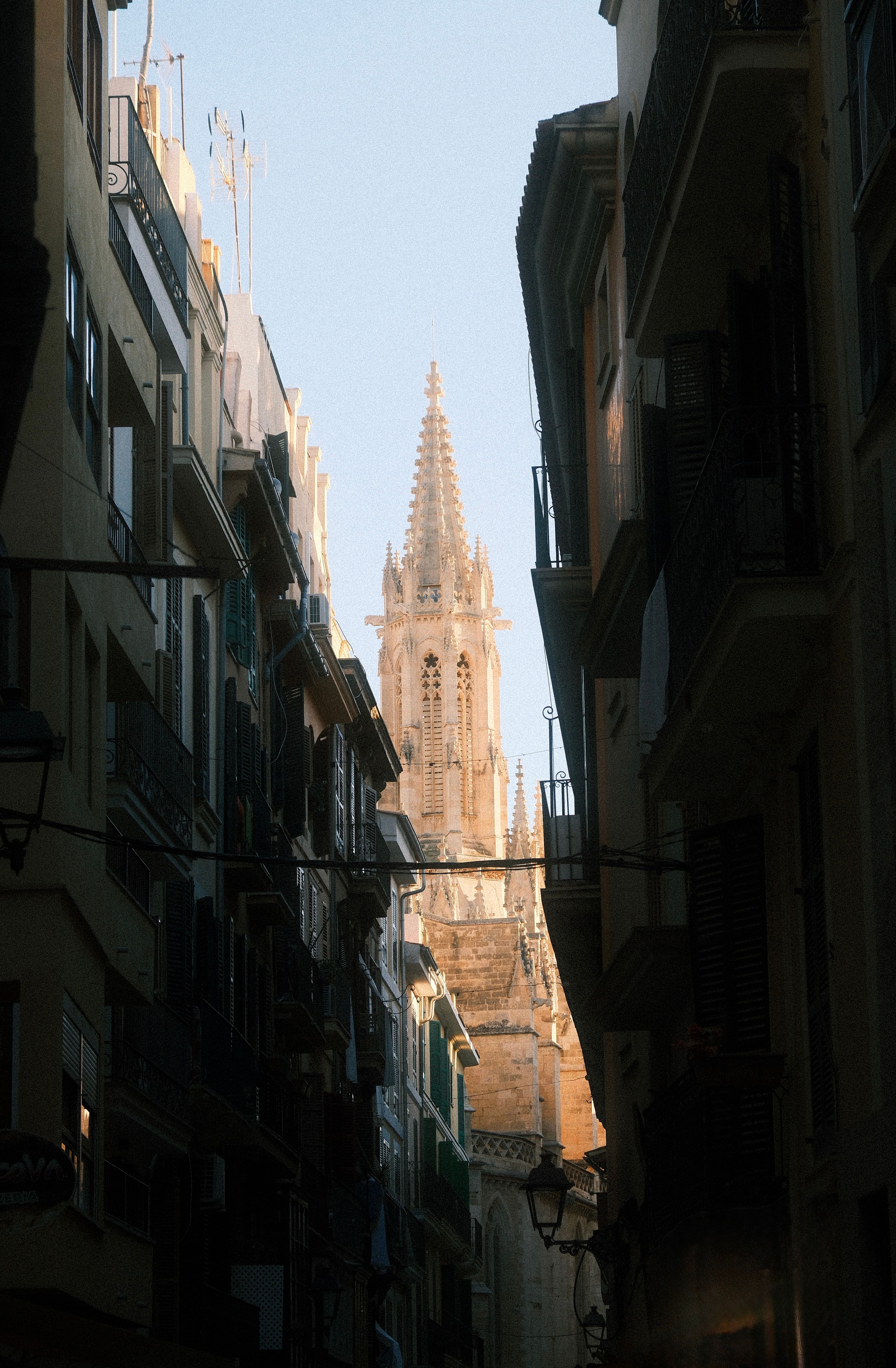 A Gothic cathedral spire illuminated by sunlight is visible between shadowy urban buildings in an alleyway.