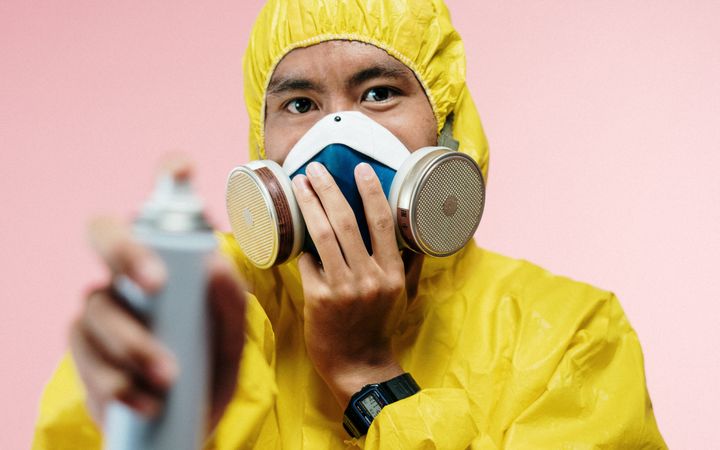 Individual wearing yellow protective suit and respirator using disinfectant spray.