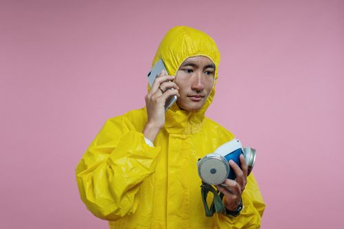 Free Person in a yellow protective suit making a phone call, holding a respirator against a pink background. Stock Photo
