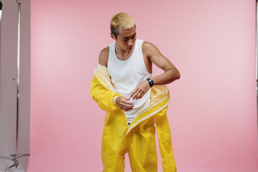 Man in a white tank top removing a yellow protective suit against a pink background.