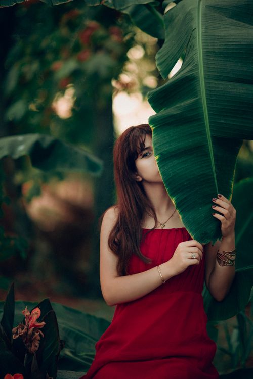 Free Elegant woman in a red dress partially hidden behind a large green leaf in a lush garden setting. Stock Photo