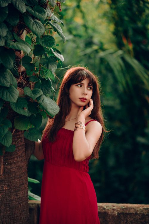 Free Portrait of a woman in a red dress surrounded by vibrant green foliage. Outdoors in natural light. Stock Photo