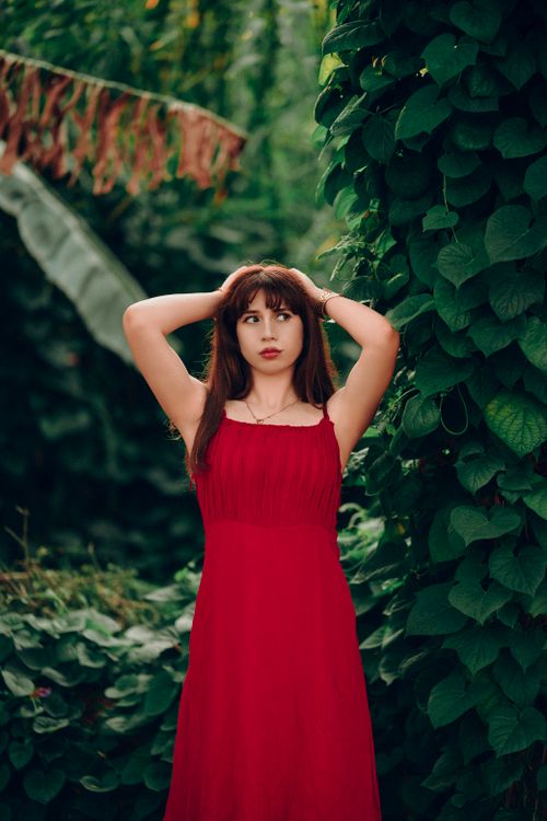 Free A woman in a red dress posing among lush green leaves, creating a striking color contrast. Stock Photo