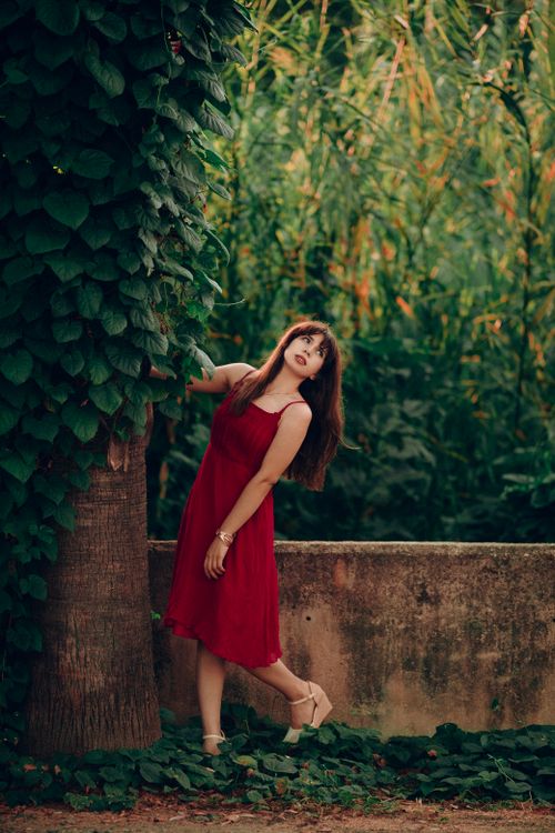 Free A woman in a red dress stands by a large tree, surrounded by lush greenery, offering a serene natural ambiance. Stock Photo